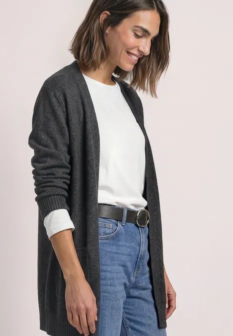 Frau in dunkelgrauer Strickjacke, weißem Shirt und blauen Jeans vor hellem Hintergrund, lächelnd seitlich stehend.