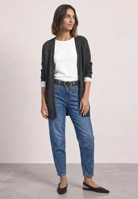 Eine Frau trägt schwarze Strickjacke, weiße Bluse, blaue Jeans und schwarze Ballerinas vor neutralem Hintergrund.
