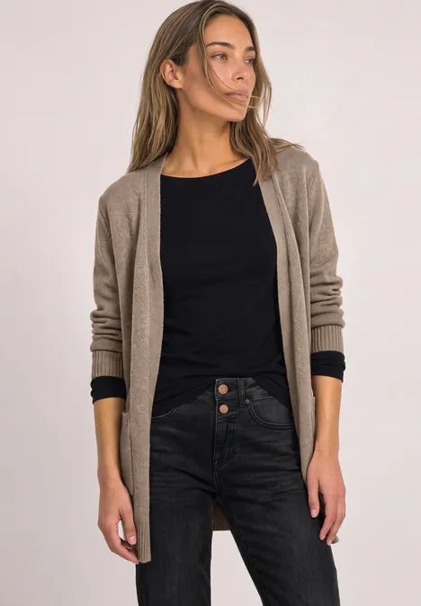Eine Frau in einem langen beigen Cardigan, schwarzem Oberteil und dunkler Jeans steht vor hellem Hintergrund.