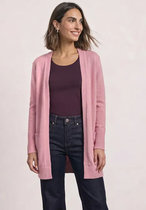 Femme portant un gilet rose, un haut bordeaux et un jean bleu foncé, souriante.
