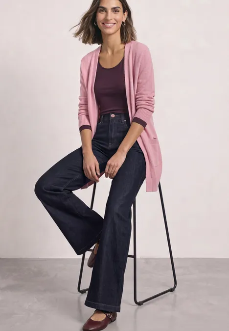 Femme assise sur un tabouret porte un cardigan rose, un haut violet et un jean flare foncé.