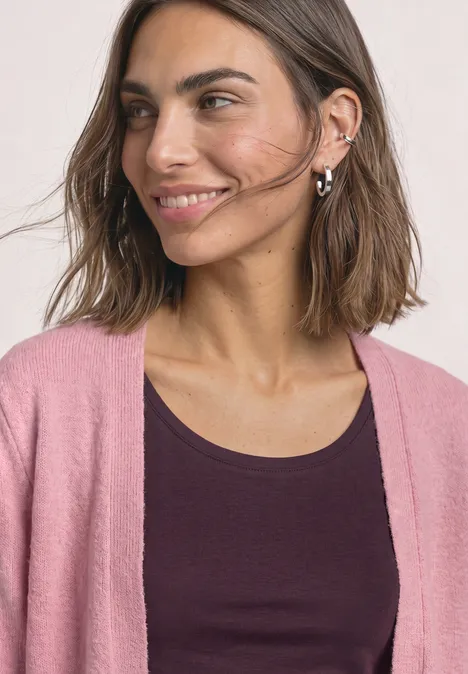 Une femme souriante aux cheveux bruns porte un haut sombre et un gilet rose.