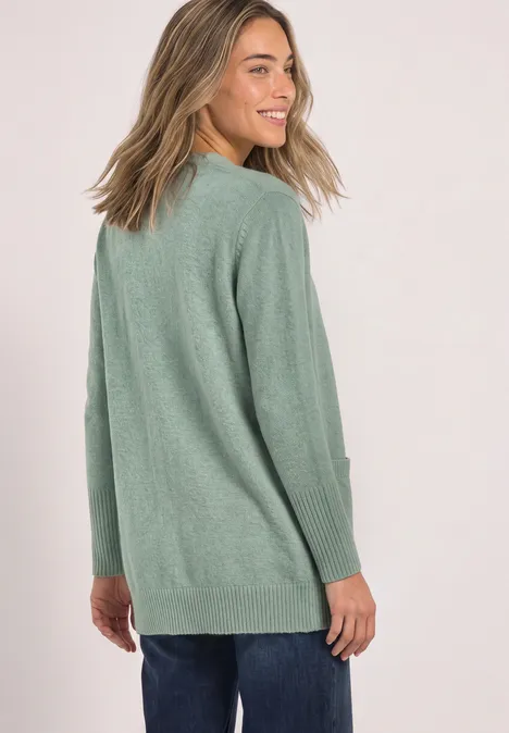 Une femme souriante aux cheveux blond-marron porte un pull vert et jette un regard par-dessus l’épaule.