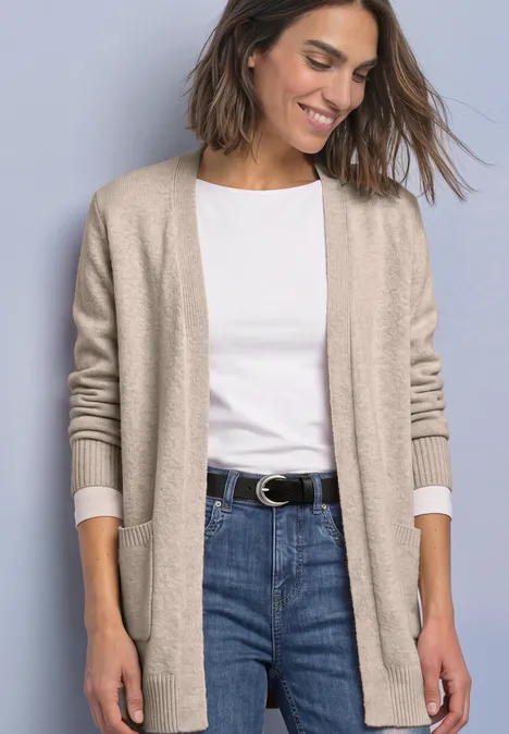 Frau in hellbeiger Strickjacke, weißem T-Shirt und Jeans vor neutralem Hintergrund