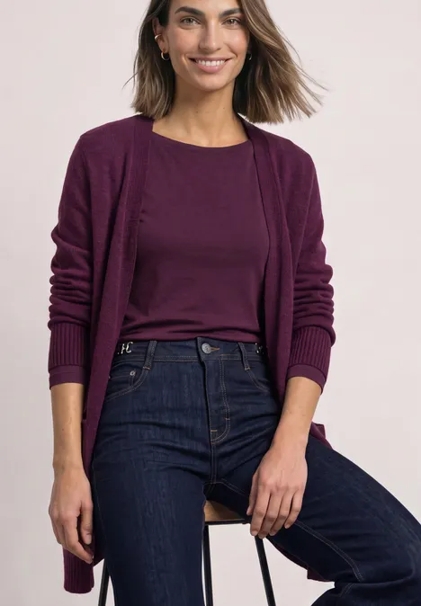 Frau mit braunen Haaren in violettem Cardigan, passendem Shirt und dunkler Jeans vor hellem Hintergrund.