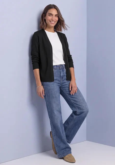 Offener Cardigan in Unifarbe Black Frau in weißem T-Shirt, schwarzer Strickjacke und weiter Jeanshose vor lila Hintergrund, lächelnd.