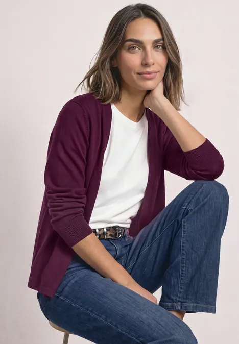 Offener Cardigan in Unifarbe berry maroon Eine lächelnde Frau sitzt, trägt ein weißes T-Shirt, violetten Cardigan, blaue Jeans und Gürtel.