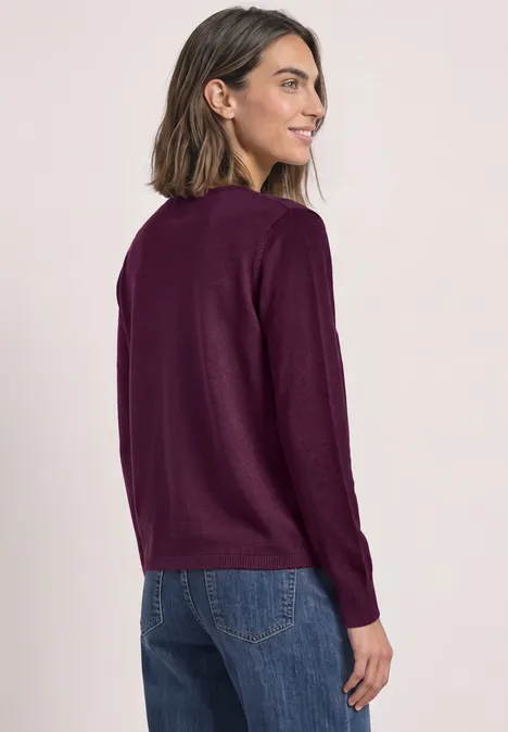 Offener Cardigan in Unifarbe berry maroon Frau trägt weichen, langärmligen, dunkellila Pullover mit Jeans, blickt über die Schulter, schlichtes Studio-Setting.