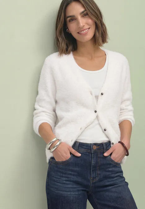 Frau in weißem Cardigan und Jeans vor hellem Hintergrund lächelnd