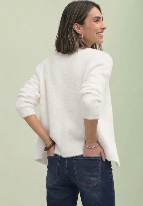 Frau mit schulterlangen Haaren trägt weißen Pullover und Jeans, lächelt seitlich in die Kamera.
