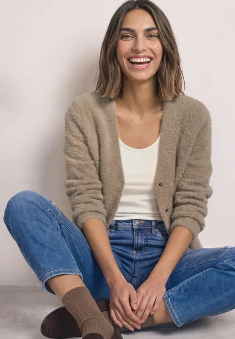 Frau sitzt lächelnd im Cardigan, Top, Jeans und Socken vor neutralem Hintergrund.
