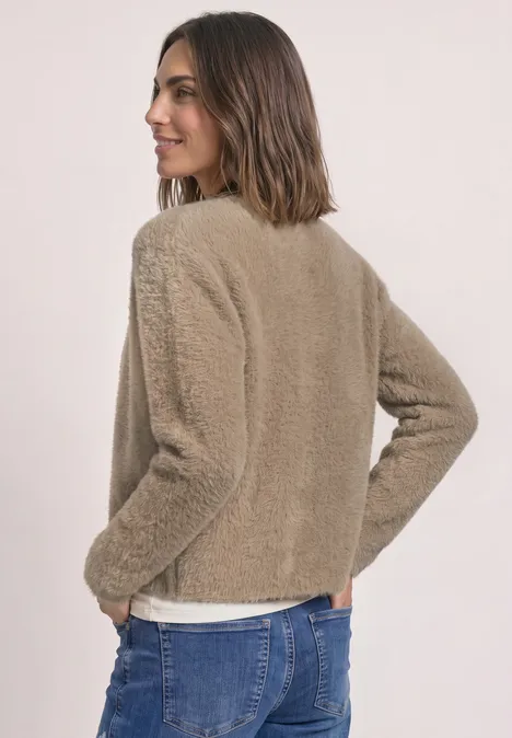 Eine Frau in beiger Teddyjacke und Jeans steht seitlich, schaut lachend über die Schulter.