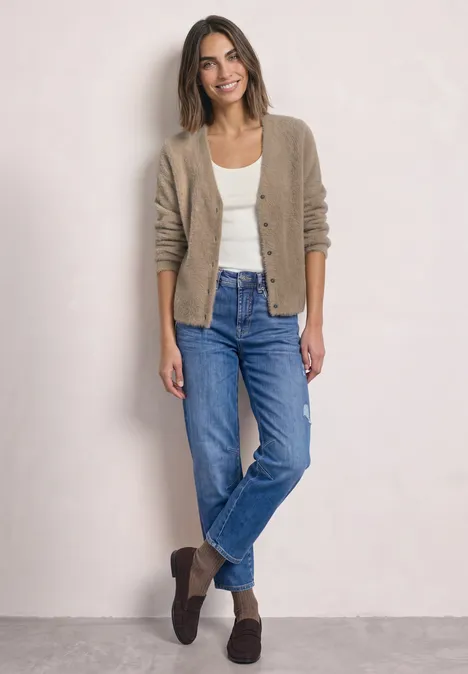 Frau in Jeans, weißem Shirt und beiger Strickjacke steht entspannt vor hellem Hintergrund.