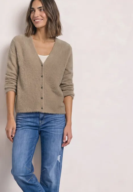 Eine Frau trägt eine beige Strickjacke und blaue Jeans, steht lächelnd vor hellem Hintergrund.