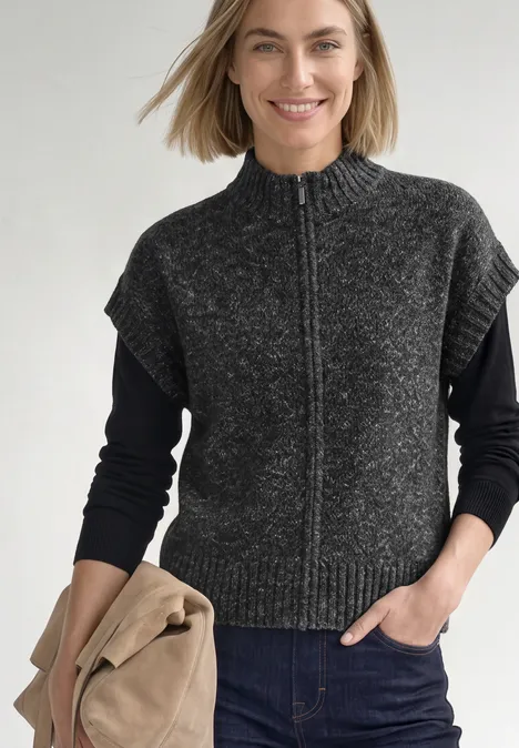 Dropped Shoulder Cardigan mit Turtle Neck und Zipper armor grey mel. Frau trägt graue, kurzärmelige Strickweste mit Reißverschluss über schwarzem Langarmshirt und Jeans.