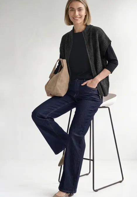 Dropped Shoulder Cardigan mit Turtle Neck und Zipper armor grey mel. Frau in legerer schwarzer Kleidung und Jeans sitzt entspannt auf hohem Hocker, lächelnd mit Tasche.