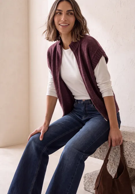 Dropped Shoulder Cardigan mit Turtle Neck und Zipper rustic rouge mel. Lächelnde Frau in weißem Shirt und dunkelblauen Jeans, mit bordeauxfarbener Jacke über den Schultern und passender Tasche.