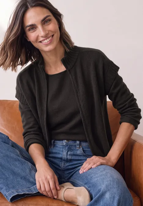 Dropped Shoulder Cardigan mit Turtle Neck und Zipper black coffee mel. Lächelnde Frau in Jeans, schwarzem Oberteil und dunklem Cardigan sitzt entspannt auf einem Sessel.