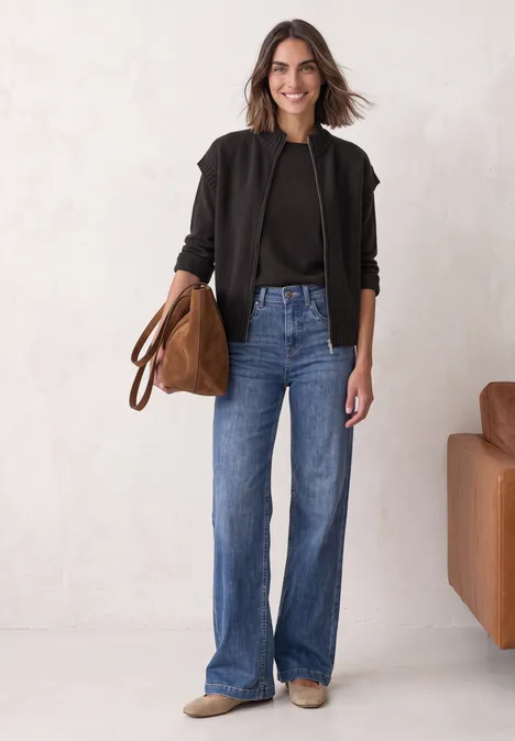 Dropped Shoulder Cardigan mit Turtle Neck und Zipper black coffee mel. Frau in schwarzer Jacke, schwarzem Top und blauen Jeans hält eine braune Tasche vor hellem Hintergrund.
