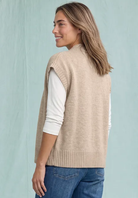 Eine Frau trägt eine beige Strickweste über weißem Langarmshirt und Jeans, blickt lächelnd zur Seite.
