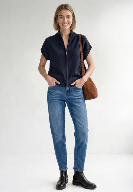 Dropped Shoulder Cardigan mit Turtle Neck und Zipper indigo night mel. Junge Frau in blauer Jeans, schwarzem Kurzarm-Oberteil und Stiefeln, mit brauner Schultertasche vor weißem Hintergrund.