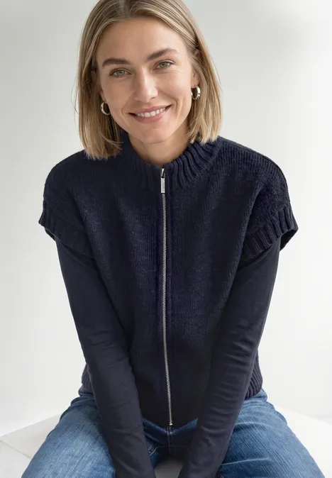 Dropped Shoulder Cardigan mit Turtle Neck und Zipper indigo night mel. Lächelnde blonde Frau sitzt in Jeans und blauem Zip-Pullover und blickt entspannt in die Kamera.