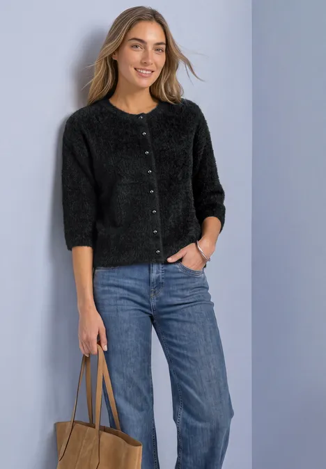 Vrouw in zwarte cardigan en blauwe spijkerbroek, met tas, glimlachend voor een neutrale achtergrond.