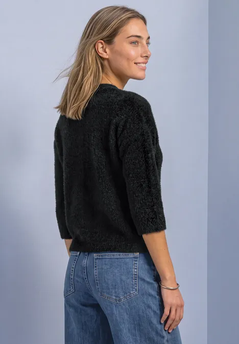 Een jonge vrouw in een zwarte, pluizige trui met driekwart mouwen en jeans kijkt glimlachend over haar schouder.