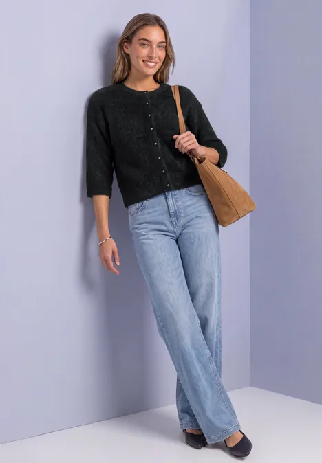 Een jonge vrouw met een zwarte cardigan en jeans, leunt tegen een lichtviolette muur en glimlacht.