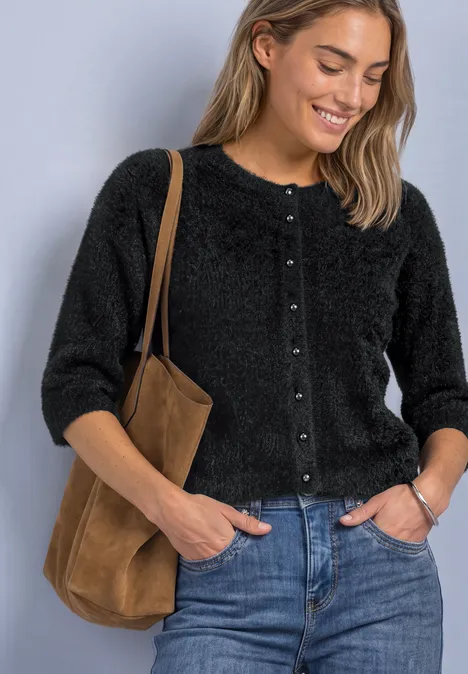 Vrouw lacht in een zwart fluffy vest, blauwe jeans en bruine schoudertas, tegen lichte achtergrond.