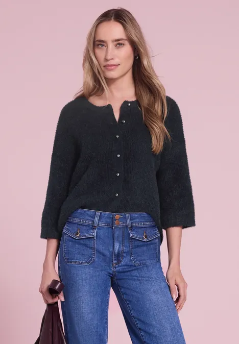 Eine Frau in schwarzer Strickjacke und blauer Jeans vor rosafarbenem Hintergrund.