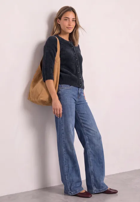 Eine junge Frau trägt weite Jeans, schwarze Strickjacke und eine braune Tasche, steht an einer Wand.