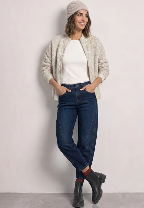 Vrouw lachend in jeans, wit t-shirt, gemêleerde cardigan, muts en laarzen, ontspannen poserend voor een lichte achtergrond.