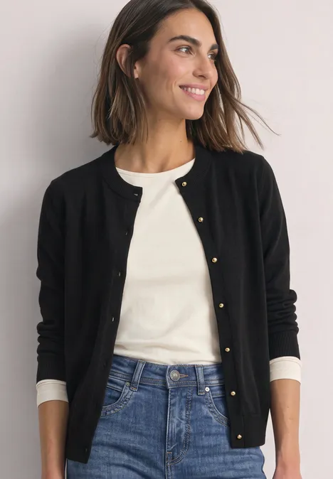 Lächelnde Frau in schwarzer Strickjacke, weißem Shirt und Jeans vor neutralem Hintergrund.