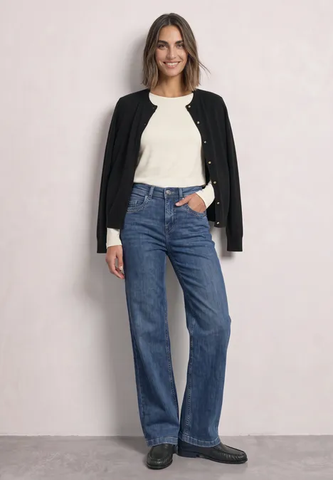 Eine Frau vor hellem Hintergrund trägt weiße Bluse, schwarze Jacke über den Schultern und gerade blaue Jeans.