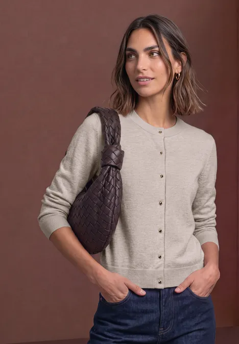 Femme en cardigan beige, jean et sac à épaule tressé brun, posant devant un fond marron.