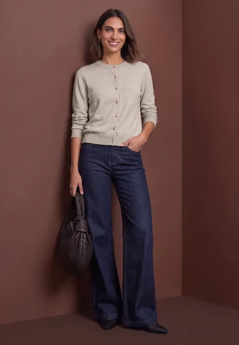 Une femme porte un gilet beige et un jean évasé bleu foncé, elle tient un sac noir.