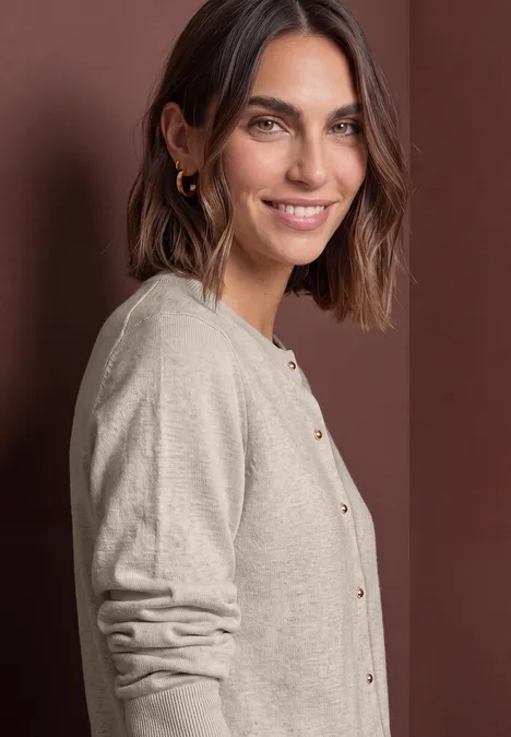 Femme souriante aux cheveux bruns mi-longs, boucles d’oreilles, gilet beige à boutons dorés devant un mur marron.