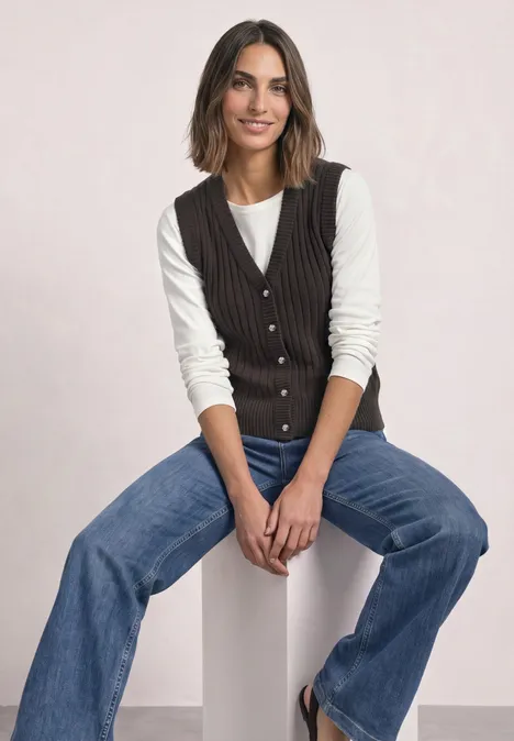 Weste mit V-Neck in Rippstruktur black coffee Eine lächelnde Frau sitzt auf einem weißen Block, trägt Jeans, weißen Pullover und dunkle Weste.