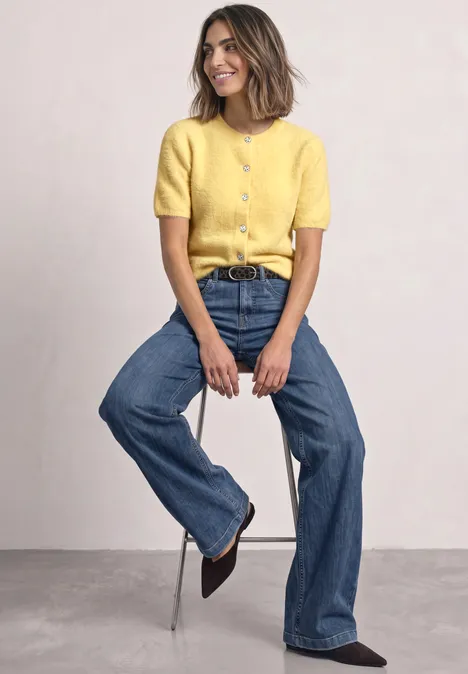 Frau trägt gelbes Oberteil und blaue Jeans und sitzt lässig auf einem Hocker.