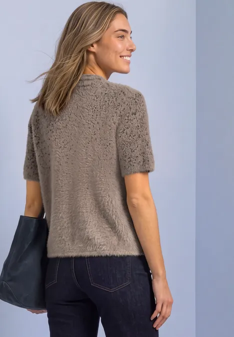 Femme portant un pull court en maille taupe, vue de dos de trois quarts, sac noir, fond clair.