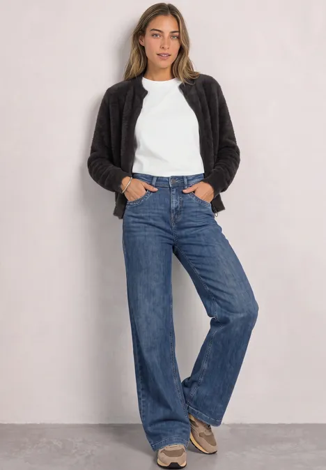 Frau in weißem T-Shirt, schwarzer Jacke und weitem Jeans lehnt lässig vor einer hellen Wand.