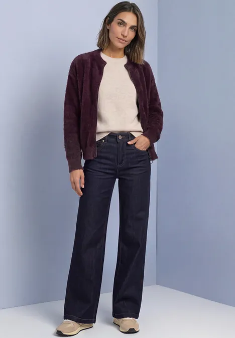 Vrouw draagt beige trui, paarse cardigan en wijd uitlopende jeans, staand voor een neutrale achtergrond.