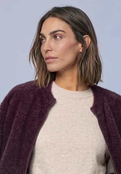 Vrouw met kort bruin haar draagt een lichte trui en paarse cardigan, kijkt opzij.