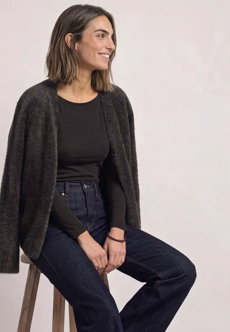 Frau sitzt lässig auf einem Hocker, trägt schwarzen Pullover, dunkle Jeans und einen lockeren Cardigan, lächelt.