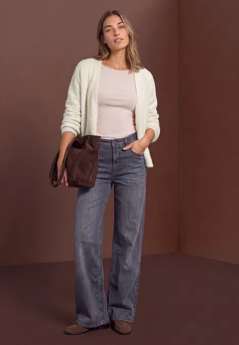 Femme en jean large, top beige et gilet blanc, portant un sac marron devant un fond brun.