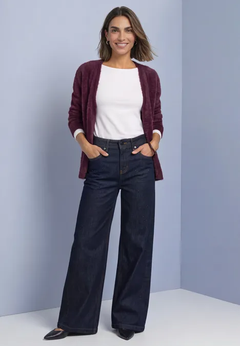 Eine Frau lächelnd in weißem Top, burgunderfarbener Strickjacke und weitem Jeans vor hellem Hintergrund.
