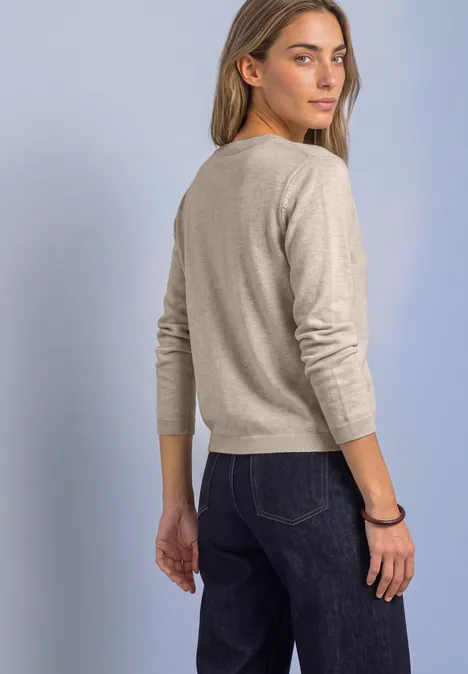Frau in beigem Pullover und dunkler Jeans dreht sich leicht zur Kamera vor hellem Hintergrund.