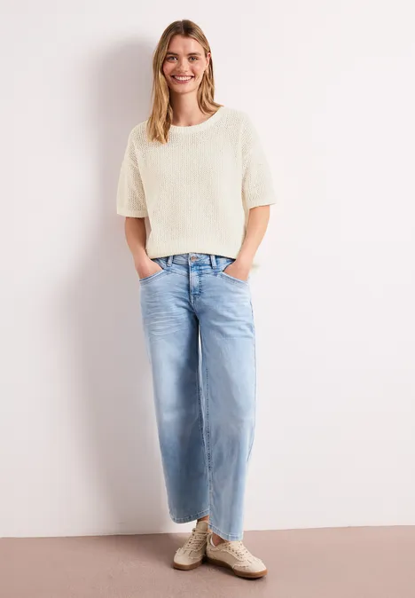 Person in cremefarbenem Kurzarm-Pullover und weiten hellblauen Jeans steht vor weißer Wand, Hände in Taschen.