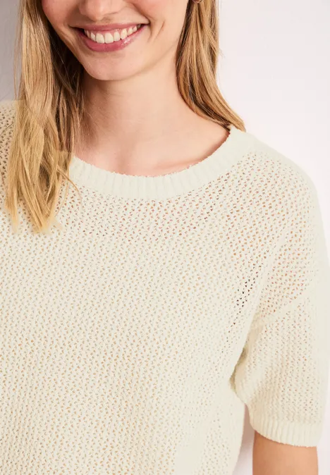 Nahaufnahme einer lächelnden Frau in einem cremefarbenen, locker gestrickten Kurzarmpullover.